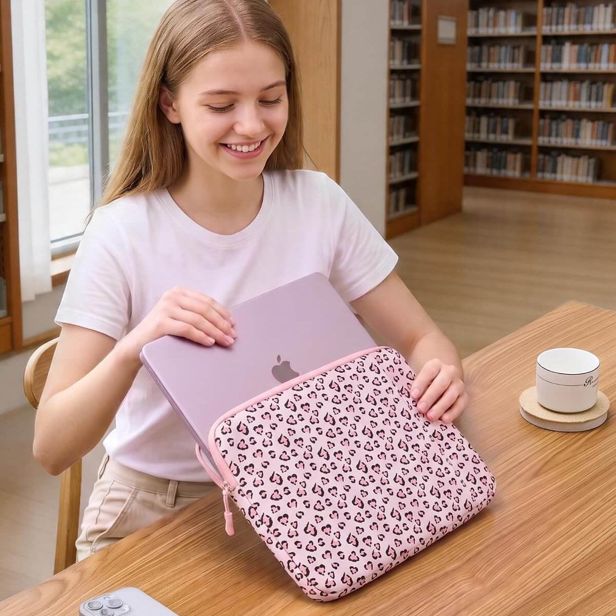A woman in a white t-shirt sits at a wooden table in a library setting, holding a light purple laptop inside a pink leopard-print laptop sleeve.