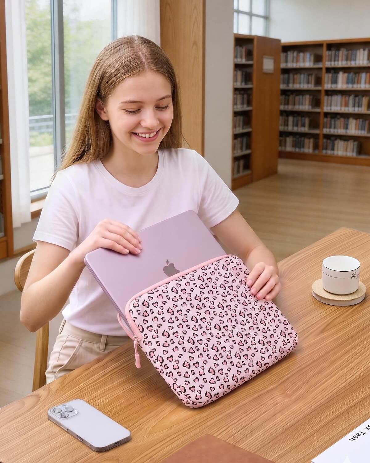 A woman in a white t-shirt sits at a wooden table in a library setting, holding a light purple laptop inside a pink leopard-print laptop sleeve.
