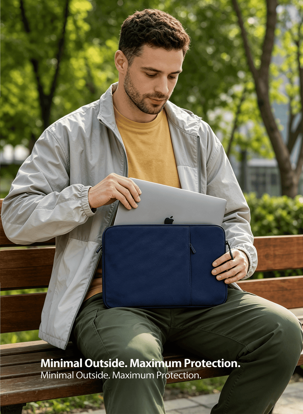 A man sitting on a park bench uses a navy blue laptop sleeve to hold a silver laptop. 