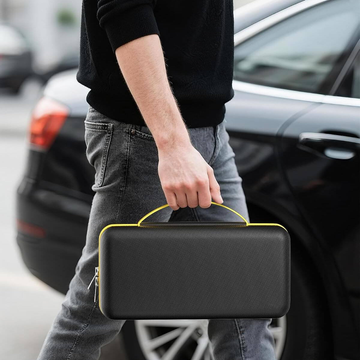 A man in a black sweater and gray jeans carries a hard case for Fanttik with a bright yellow handle and zipper trim, walking past a black car outdoors.