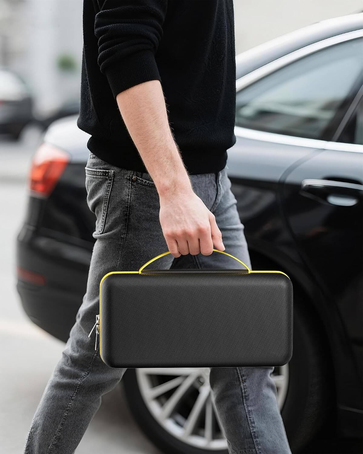 A man in a black sweater and gray jeans carries a hard case for Fanttik with a bright yellow handle and zipper trim, walking past a black car outdoors.