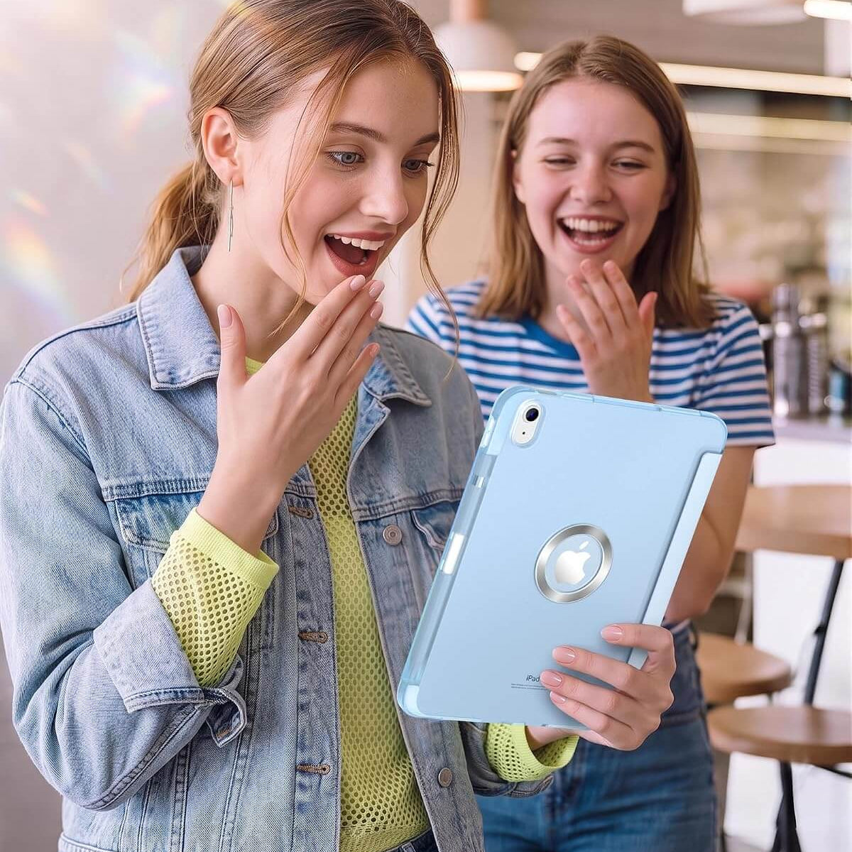 Two young women in a bright cafe setting are excitedly looking at a light blue iPad A16 11th/10th Gen Smart Case. The woman in the foreground is wearing a light-wash denim jacket over a yellow-green top, and the woman behind her is wearing a blue and white striped shirt.