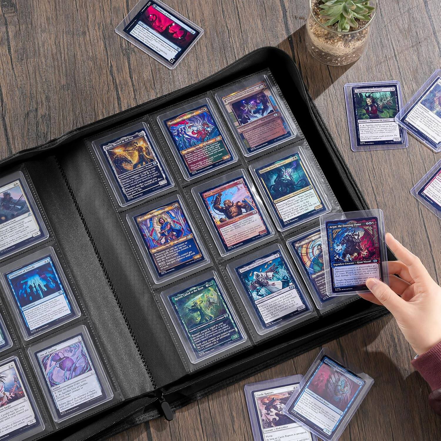 A black card binder filled with collectible trading cards, shown open on a wooden table. A hand is holding one card, and several cards and a small potted succulent are placed around it.