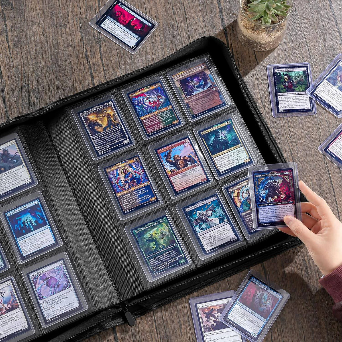 A black card binder filled with collectible trading cards, shown open on a wooden table. A hand is holding one card, and several cards and a small potted succulent are placed around it.