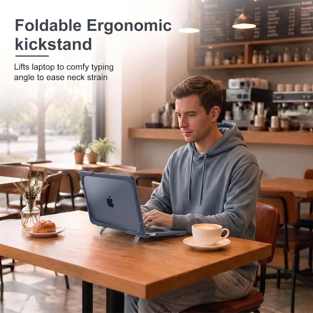 A man in a gray hoodie sits at a wooden cafe table using a macbook on a foldable ergonomic kickstand. The scene includes text overlay reading 'Foldable Ergonomic kickstand' and shows the stand lifting the laptop to a comfortable typing angle.