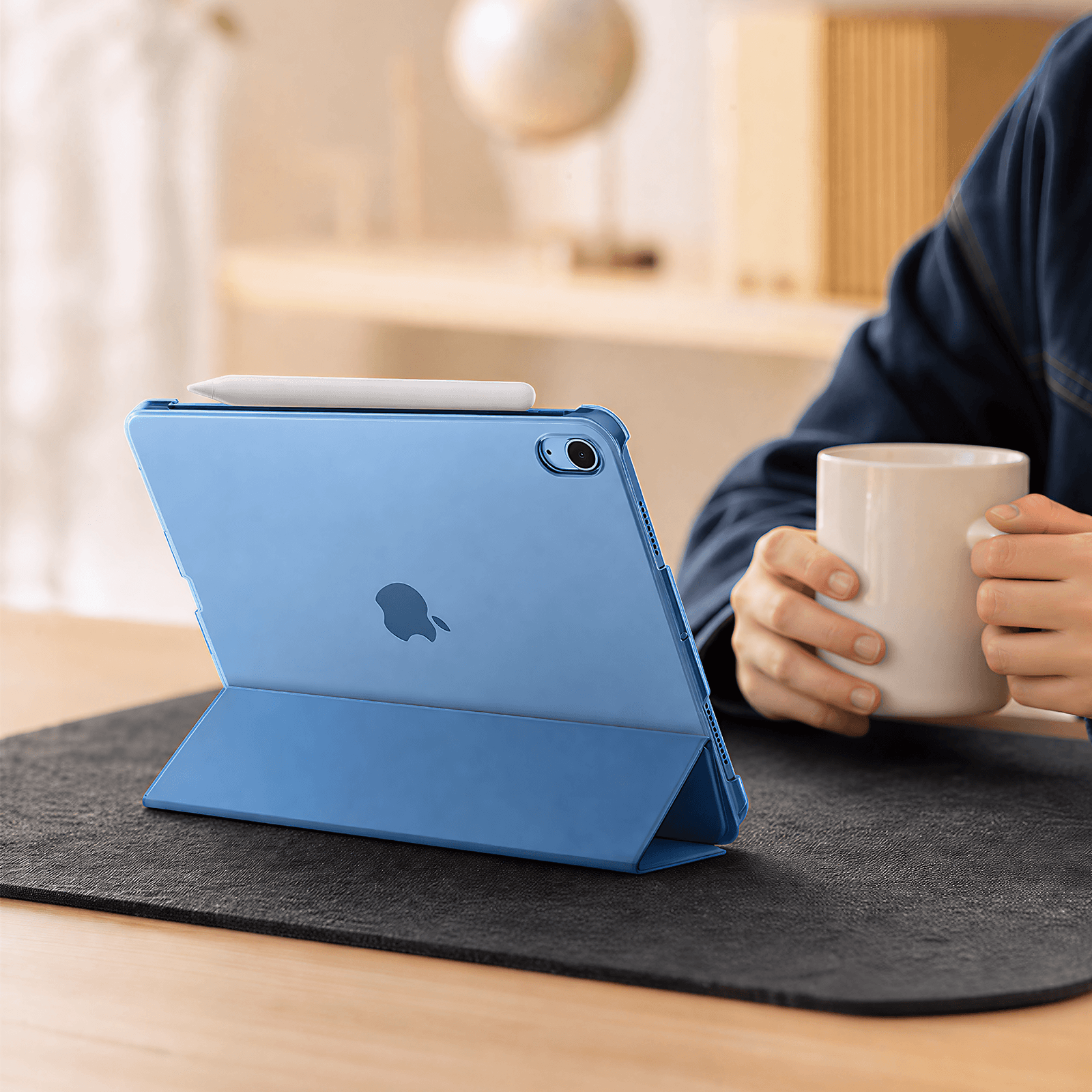 A blue iPad case with a built-in kickstand is shown on a desk, with a person holding a white mug in the background.