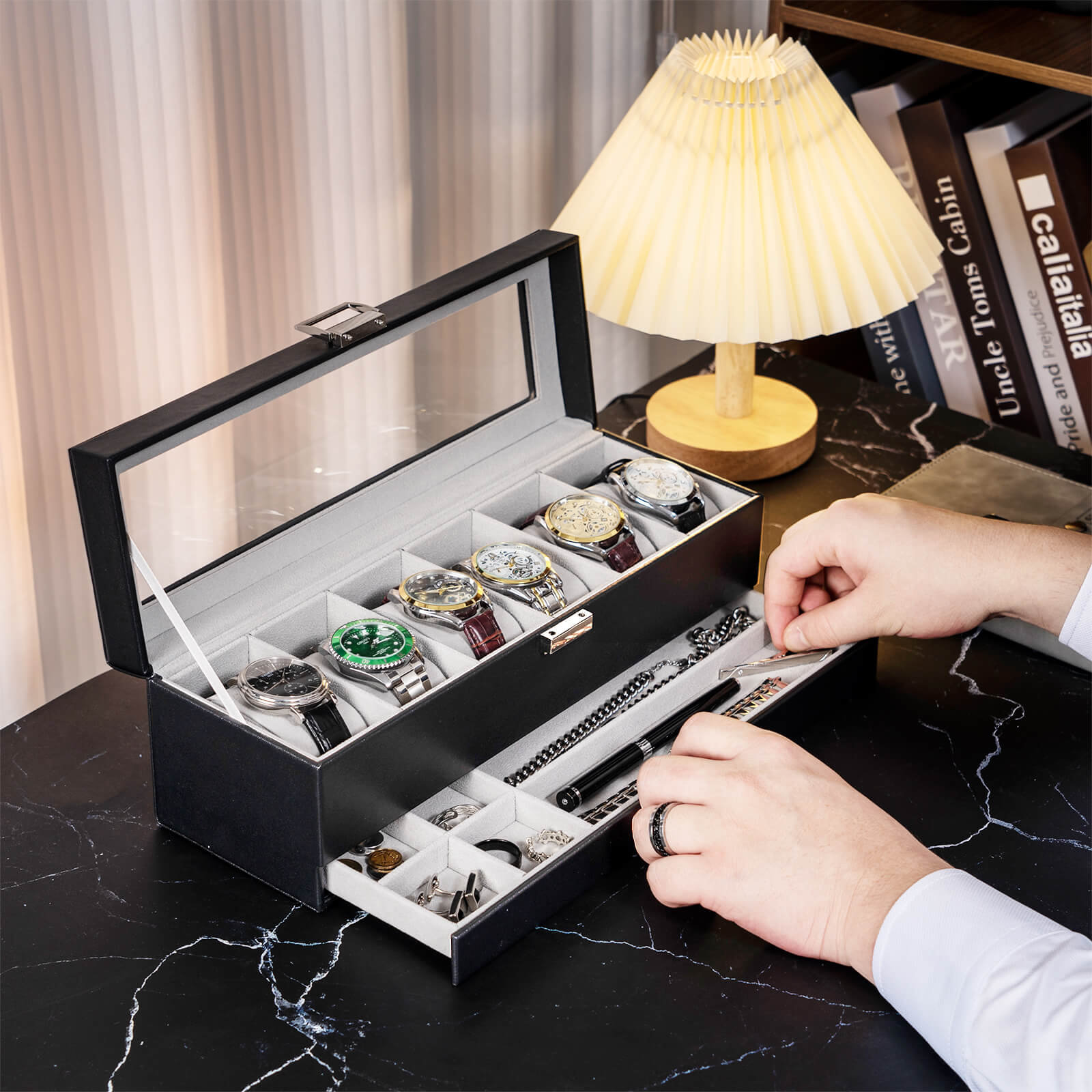 Black jewelry box with watches and jewelry on a marble surface with a lamp in the background.