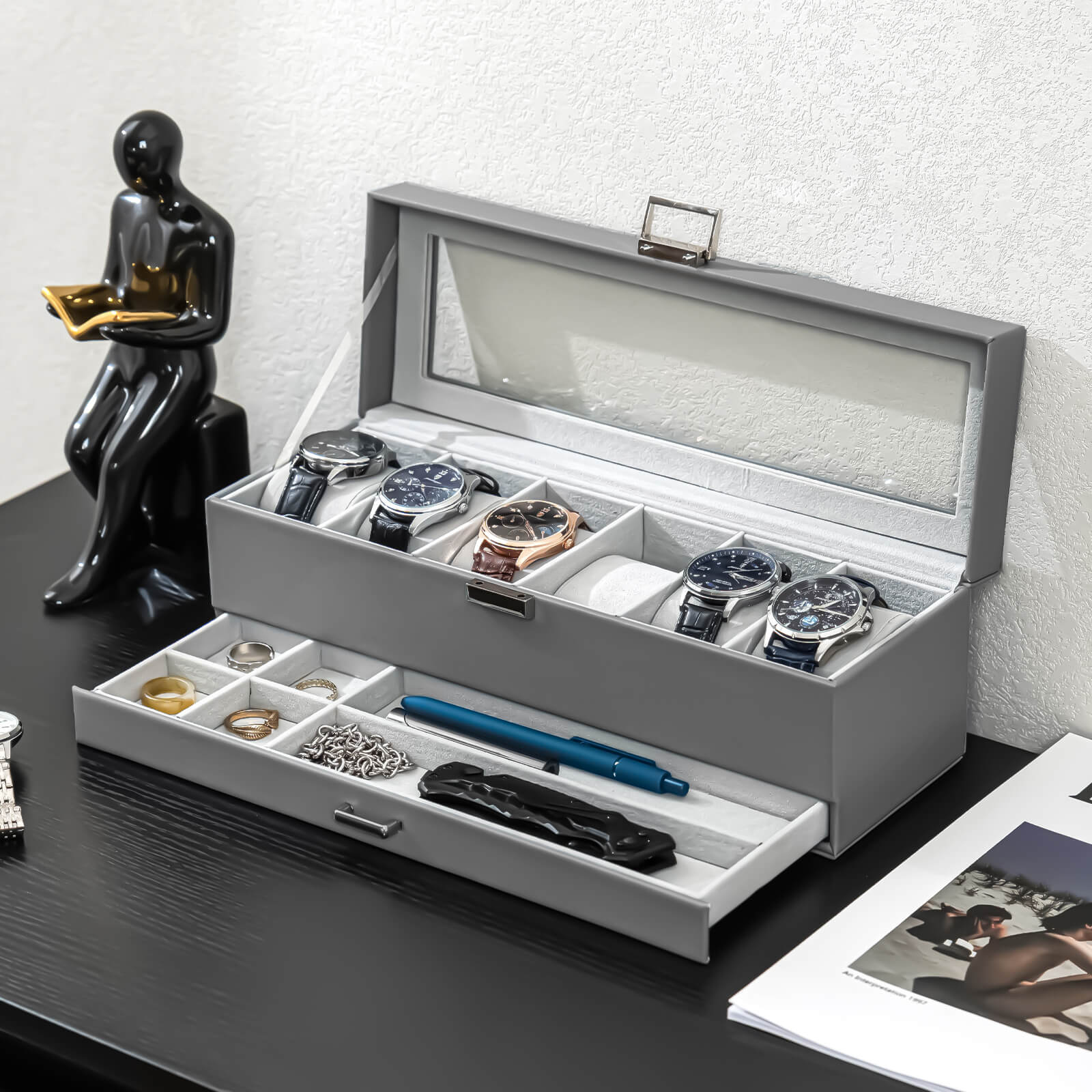 Gray jewelry and watch box with watches, rings, and a pen on a dark surface.