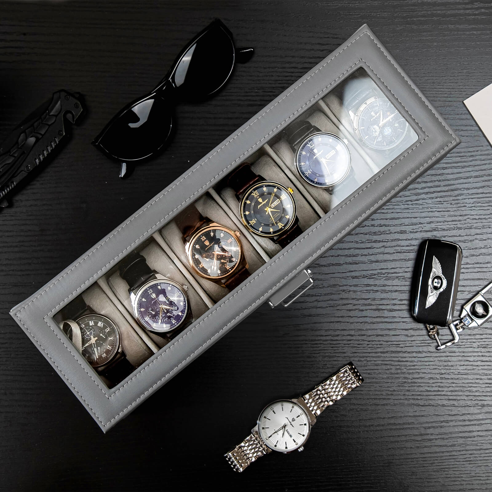 Gray watch box with six watches on a dark surface with sunglasses and a car keychain.