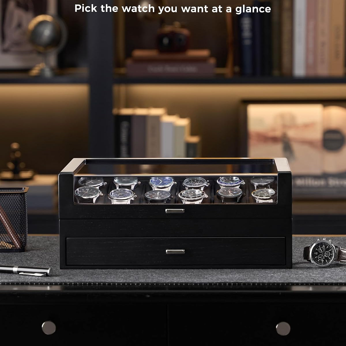 A black watch display box with a clear glass top window showing several wristwatches inside, placed on a dark desk in a dimly lit home office setting. 