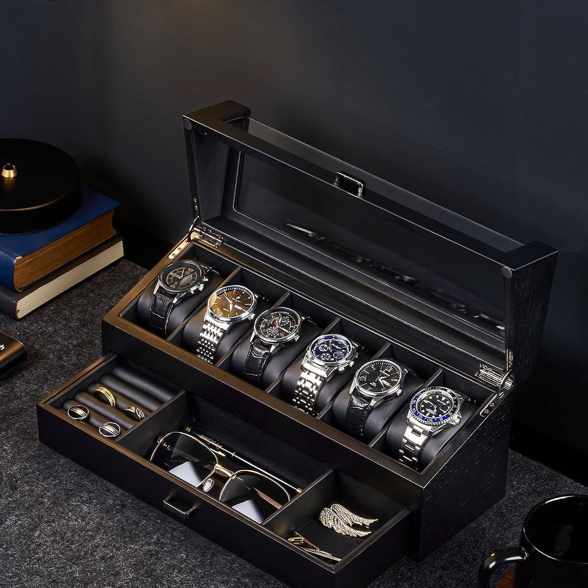 A black watch and accessory storage box with a glass top lid, shown open on a dark tabletop. 