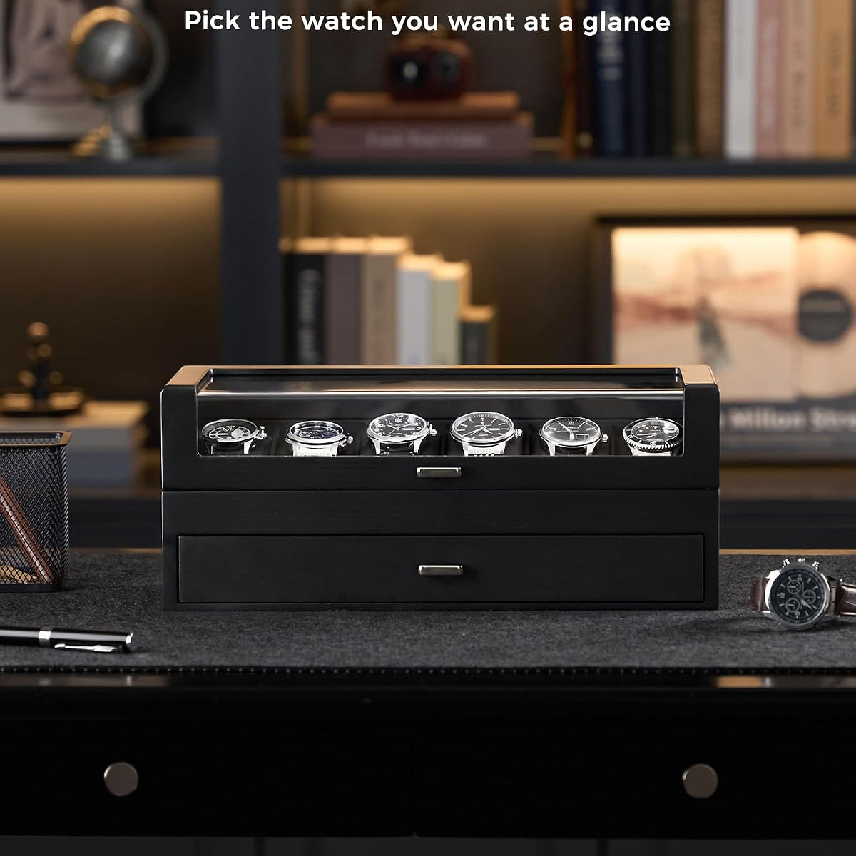 A black watch winder box with six watch slots on top and a lower drawer, shown on a dark desk in a home office setting with shelves in the background. 