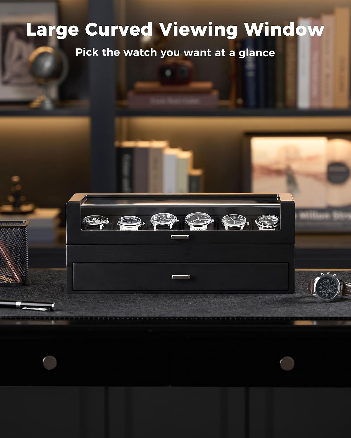 A black watch winder box with six watch slots on top and a lower drawer, shown on a dark desk in a home office setting with shelves in the background. 