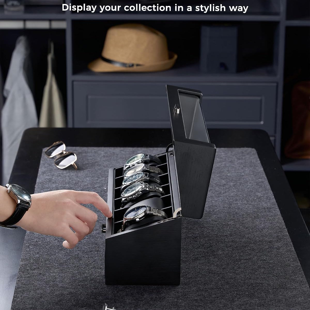A black rectangular watch display box with an open lid and multiple watch slots, shown on a dark tabletop in a room setting.