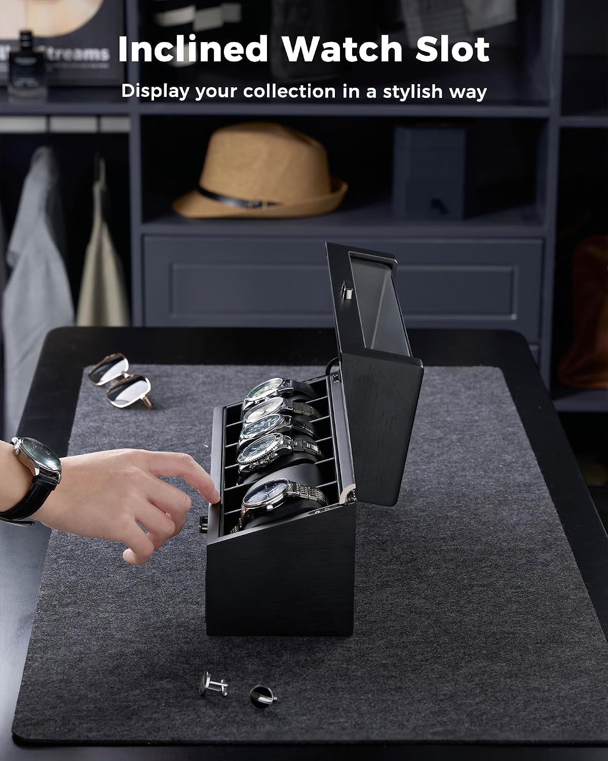 A black rectangular watch display box with an open lid and multiple watch slots, shown on a dark tabletop in a room setting.