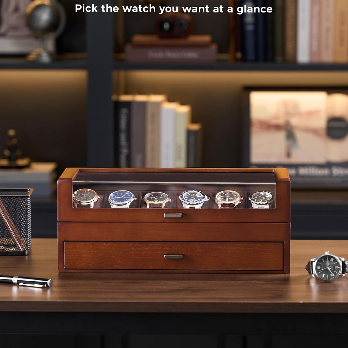 A wooden watch display box with a glass top window showing several watches inside, placed on a desk in a blurred office setting. 