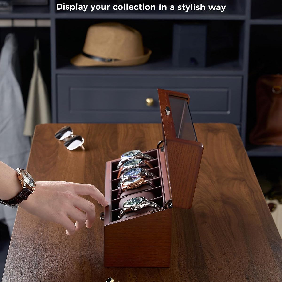A wooden watch display box with an open lid and angled watch slots, shown on a wooden table in a styled interior. 