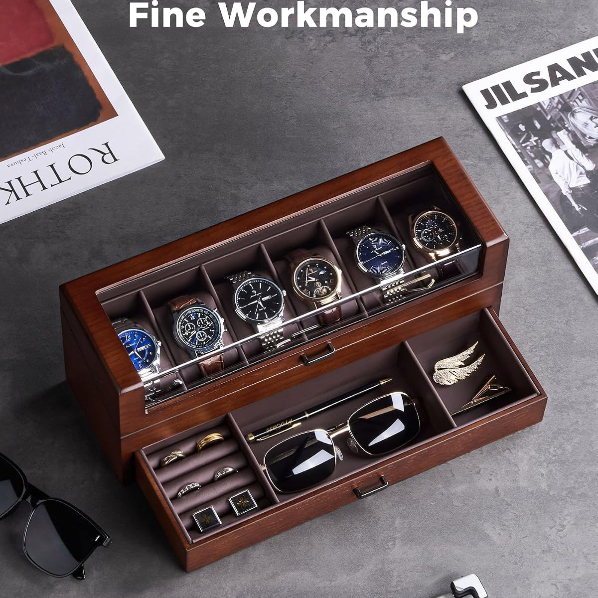 A premium wooden watch and accessory organizer with a top watch display and an open drawer holding sunglasses, cufflinks, and a tie, styled on a gray surface with magazines and a laptop nearby. 