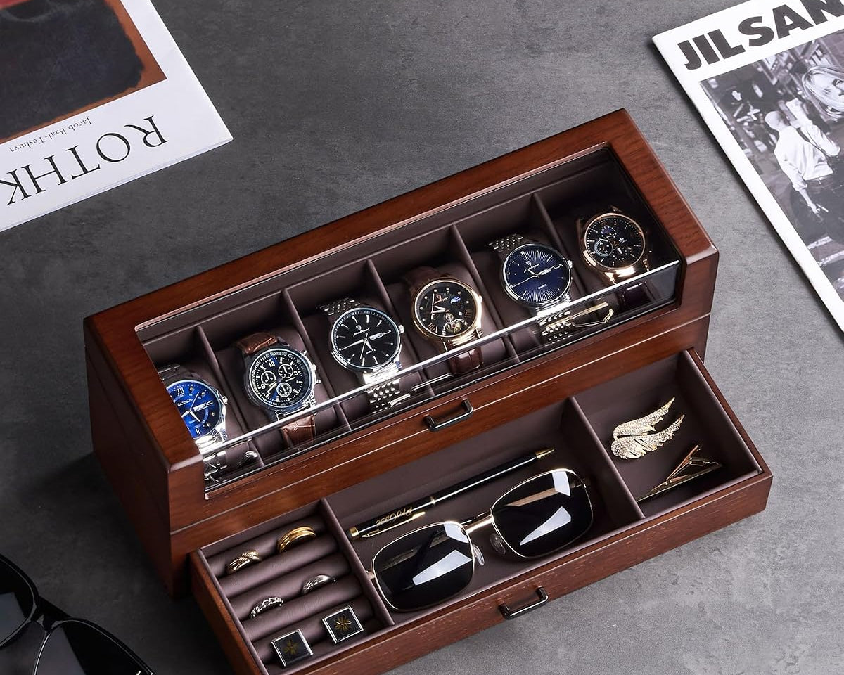 A premium wooden watch and accessory organizer with a top watch display and an open drawer holding sunglasses, cufflinks, and a tie, styled on a gray surface with magazines and a laptop nearby. 