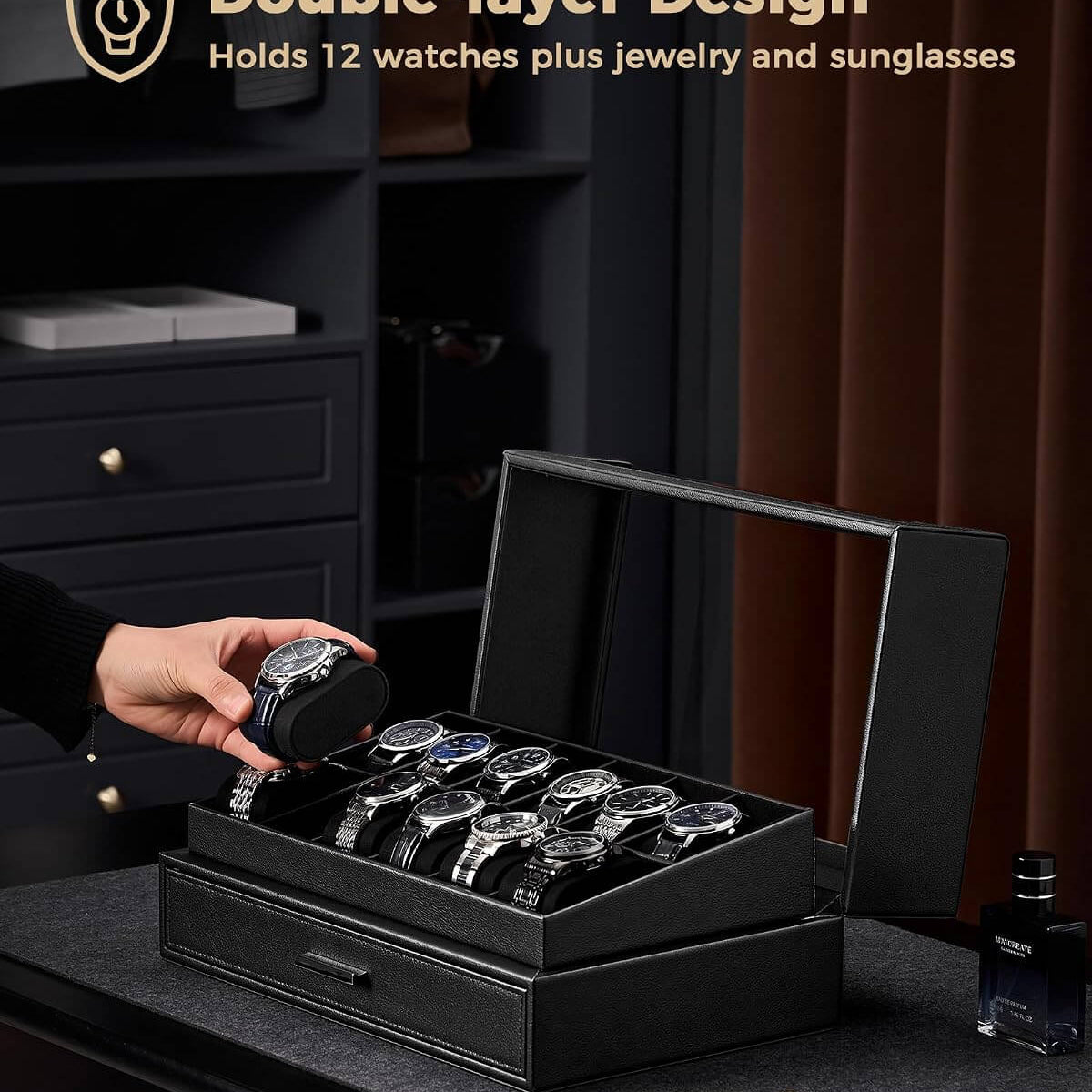 A black double-layer watch storage box with a clear  lid is shown open on a dark table, holding several watches and jewelry. A hand is placing a watch into the box.