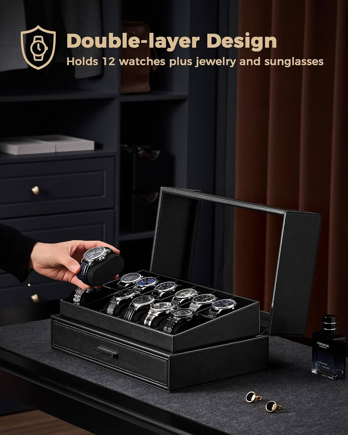 A black double-layer watch storage box with a clear  lid is shown open on a dark table, holding several watches and jewelry. A hand is placing a watch into the box.