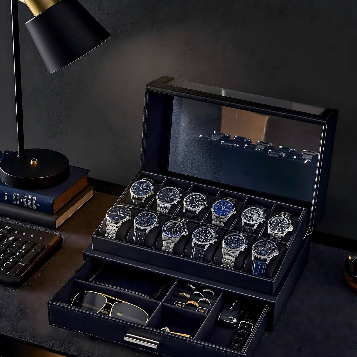 A luxury watch display case and organizer set on a dark desk, featuring a large open navy blue case with multiple watch slots holding silver and blue-faced watches, a lower drawer with accessories, and a black desk lamp beside it.
