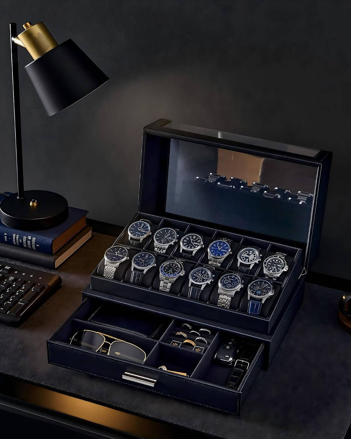 A luxury watch display case and organizer set on a dark desk, featuring a large open navy blue case with multiple watch slots holding silver and blue-faced watches, a lower drawer with accessories, and a black desk lamp beside it.