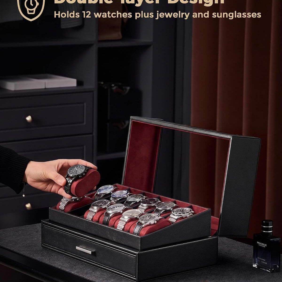 A black double-layer watch storage box with a red interior is shown open on a dark table, holding several watches and jewelry. A hand is placing a watch into the top compartment.