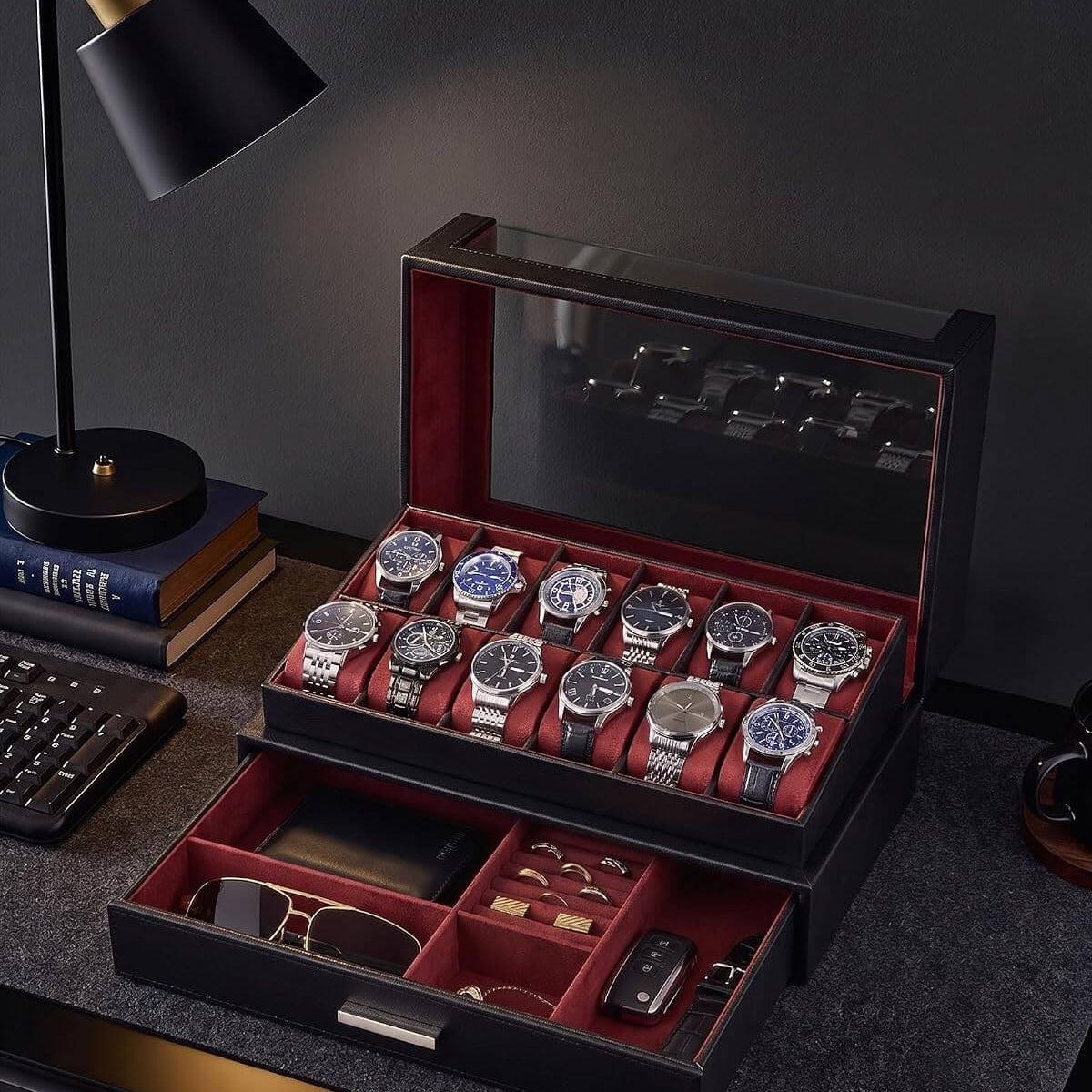 A black watch storage box with a glass top lid and an open lower drawer, filled with multiple wristwatches, sunglasses, and accessories. The box has a red interior and sits on a dark desk next to a black desk lamp and books.