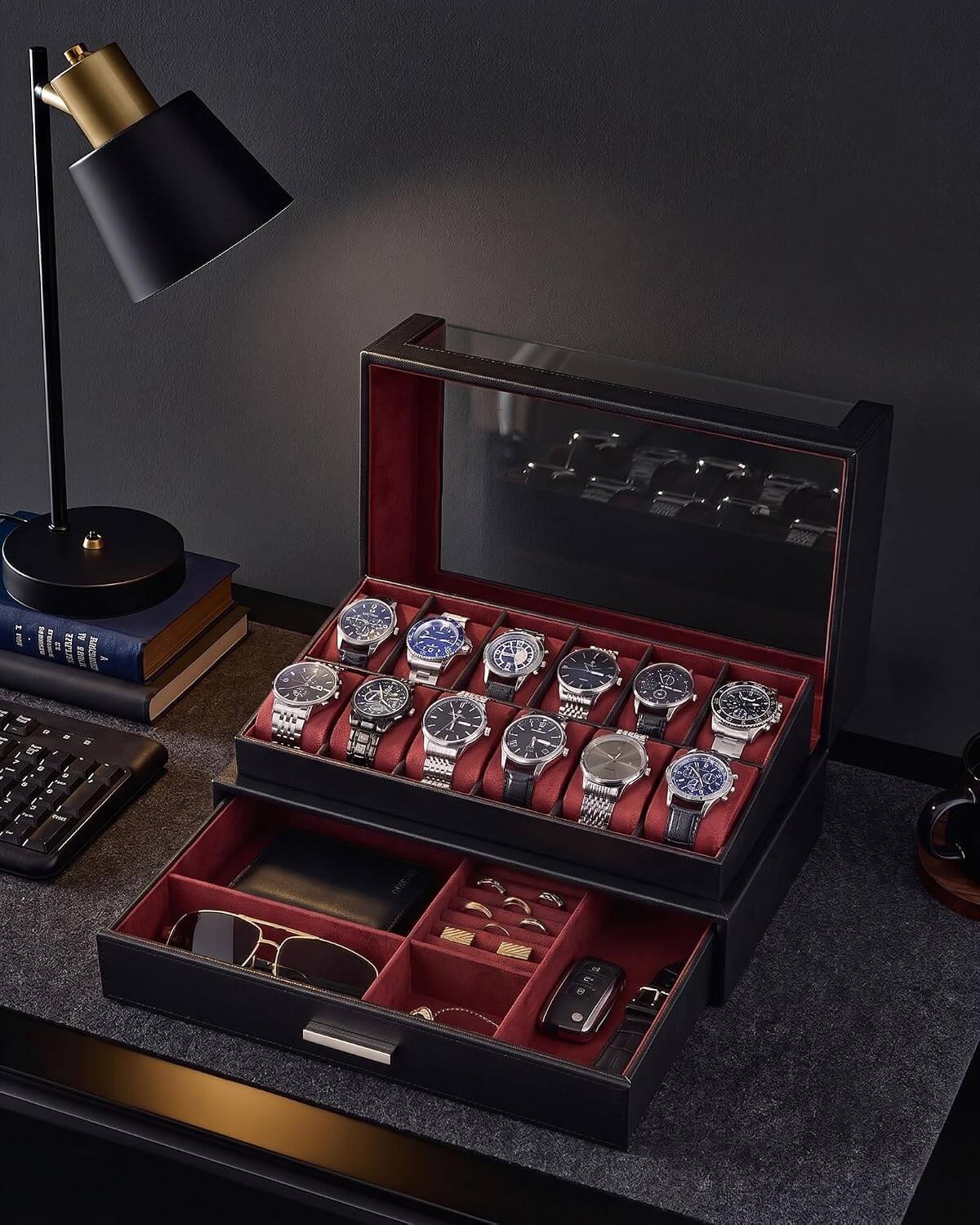 A black watch storage box with a glass top lid and an open lower drawer, filled with multiple wristwatches, sunglasses, and accessories. The box has a red interior and sits on a dark desk next to a black desk lamp and books.
