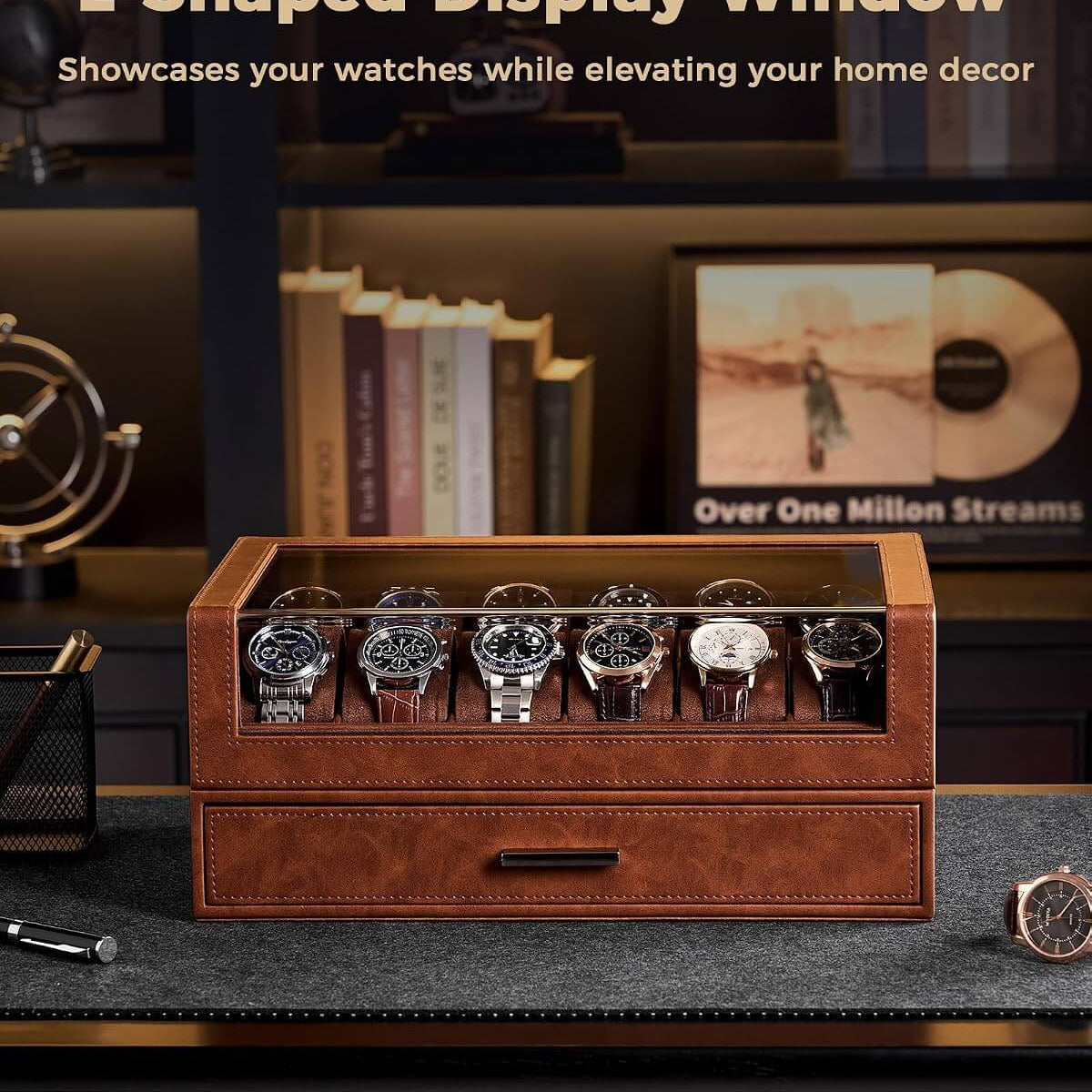 A brown leather watch display box with a glass top window, shown holding multiple wristwatches on a dark desk. The scene is styled in a home office setting with books, a record player, and shelves in the background