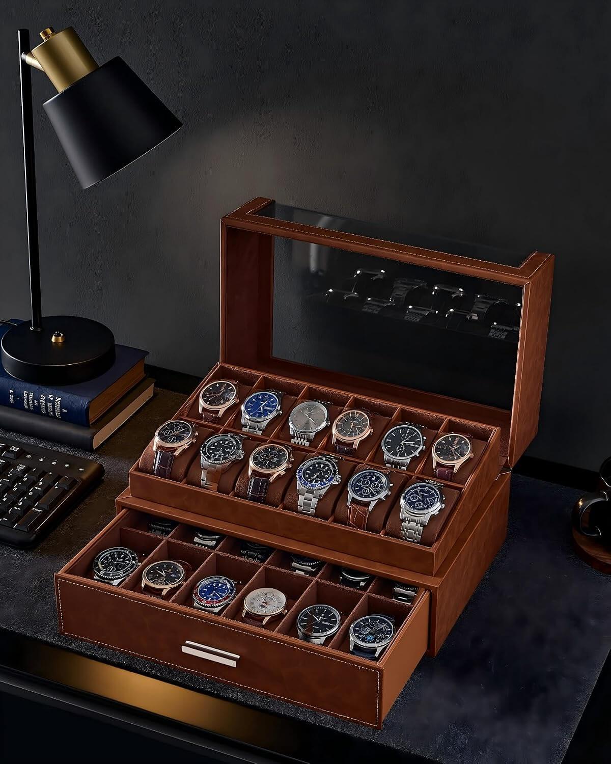 A brown watch display case with a glass top, shown open on a dark desk. The case contains multiple rows of wristwatches, and a matching drawer sits below.
