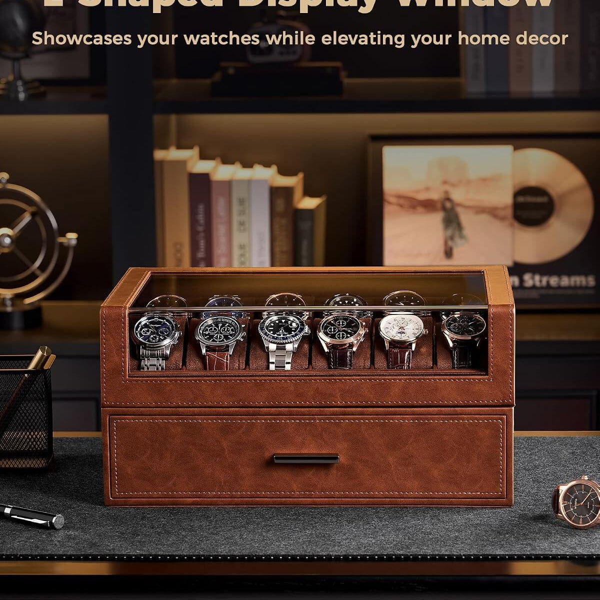 A brown leather watch display drawer with a glass top window showing several watches inside, placed on a dark desk in a styled home office setting. 