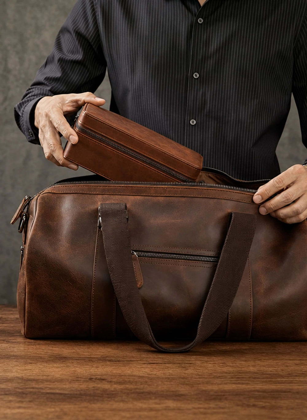 A brown leather duffel bag on a wooden table, with a person in a dark striped shirt holding a matching brown leather case inside the open bag.