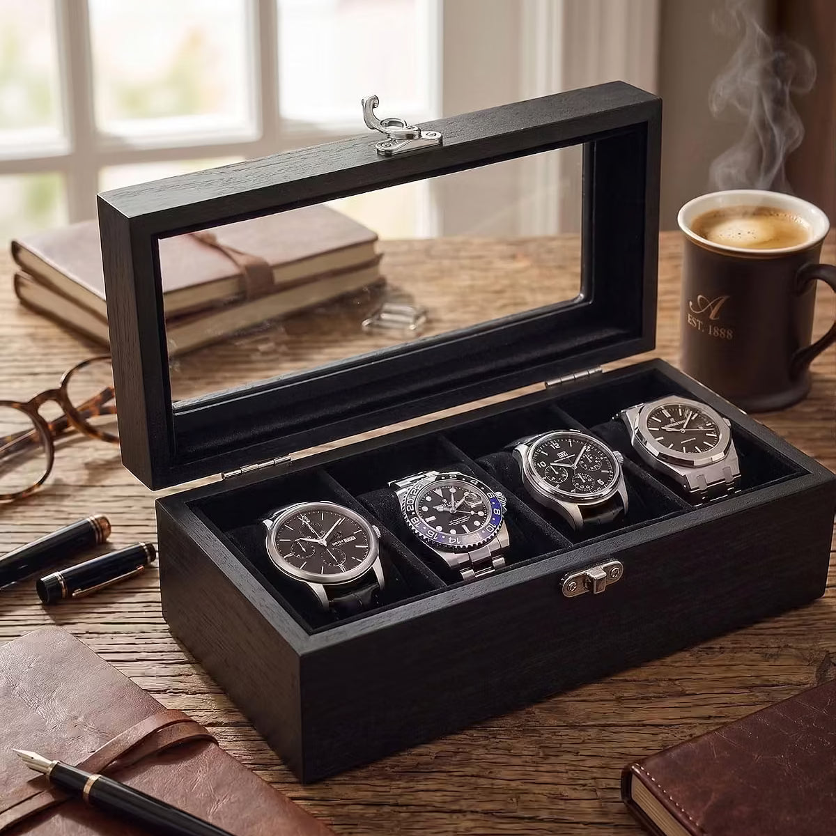 A black wooden watch box with a glass lid, shown open on a wooden table with four watches inside. The scene includes a coffee mug, notebooks, and eyeglasses in a warm, styled interior setting.