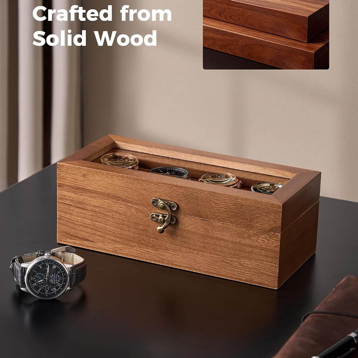A rectangular wooden watch box with a transparent top, shown on a dark tabletop in a styled indoor setting. Several watches are stored inside, and a small inset at the top right shows a close-up of the wood grain.
