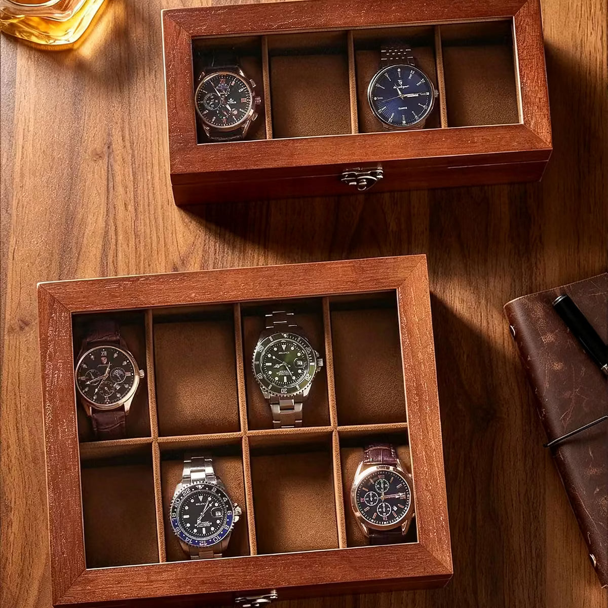Two wooden watch display boxes with glass lids are arranged on a wooden table, each holding four wristwatches. A glass of amber liquid and part of a brown notebook are visible as styled props.