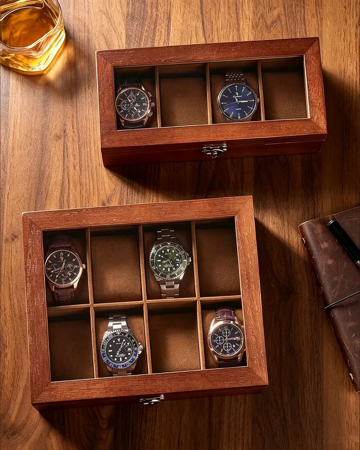 Two wooden watch display boxes with glass lids are arranged on a wooden table, each holding four wristwatches. A glass of amber liquid and part of a brown notebook are visible as styled props.
