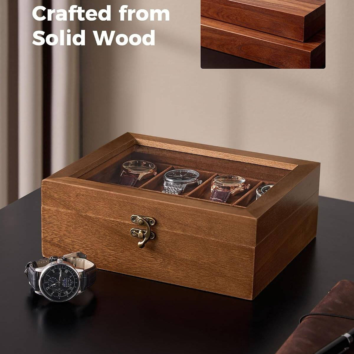 A wooden watch display box with a glass top, shown on a dark tabletop in a styled interior setting. The box has a warm medium-brown wood finish, a small metal latch on the front, and multiple watch slots containing watches.