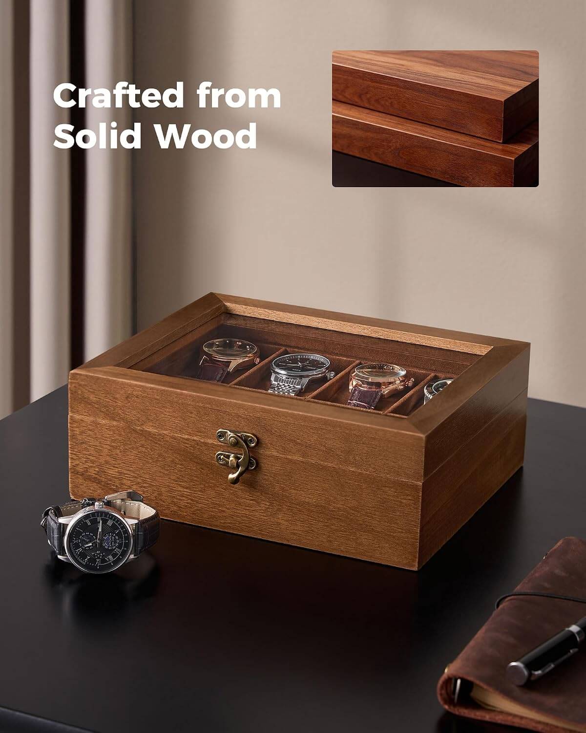 A wooden watch display box with a glass top, shown on a dark tabletop in a styled interior setting. The box has a warm medium-brown wood finish, a small metal latch on the front, and multiple watch slots containing watches.
