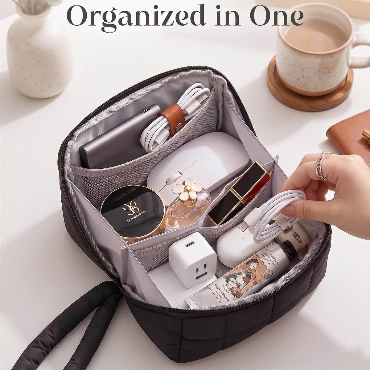 A black puffy tech pouch shown open on a white tabletop, filled with various electronics, cosmetics, and accessories. A hand is reaching toward the bag, and a coffee cup and vase are visible in the background.