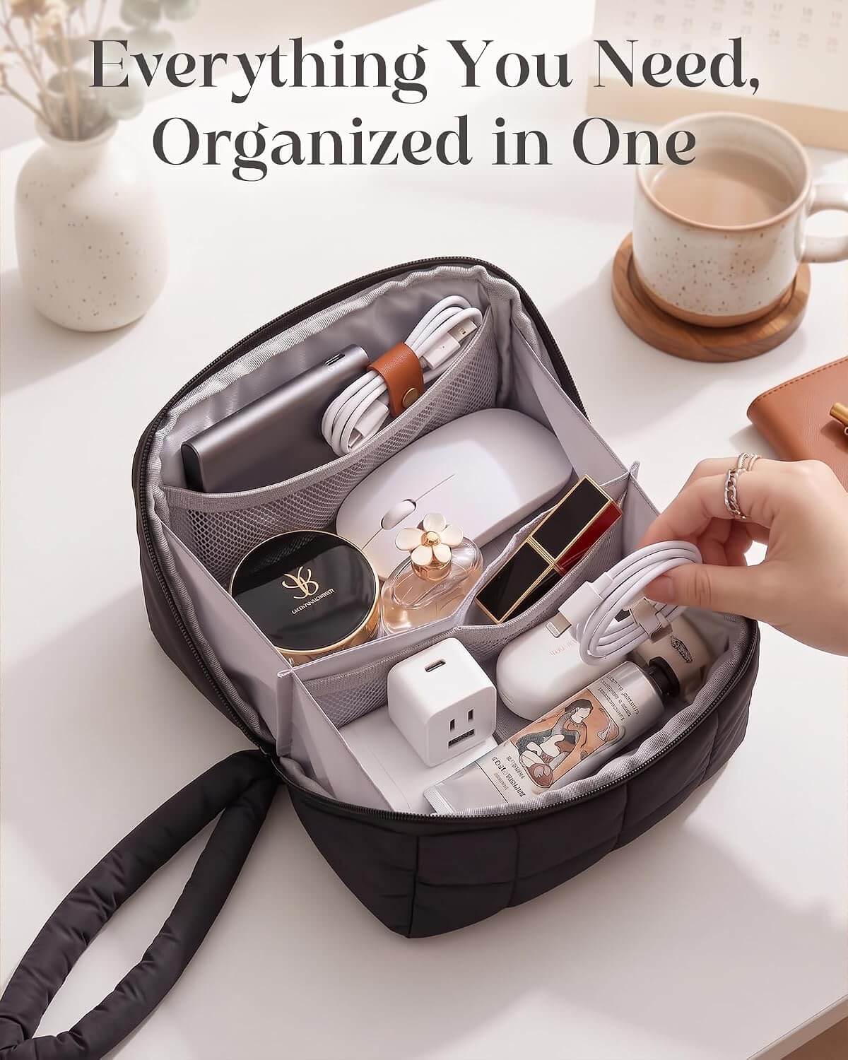 A black puffy tech pouch shown open on a white tabletop, filled with various electronics, cosmetics, and accessories. A hand is reaching toward the bag, and a coffee cup and vase are visible in the background.