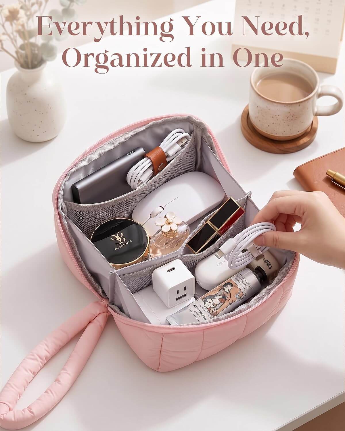 A pink puffy tech pouch with multiple interior compartments, shown open on a white desk with various personal items inside and a hand reaching in. The text at the top reads, 'Everything You Need, Organized in One.