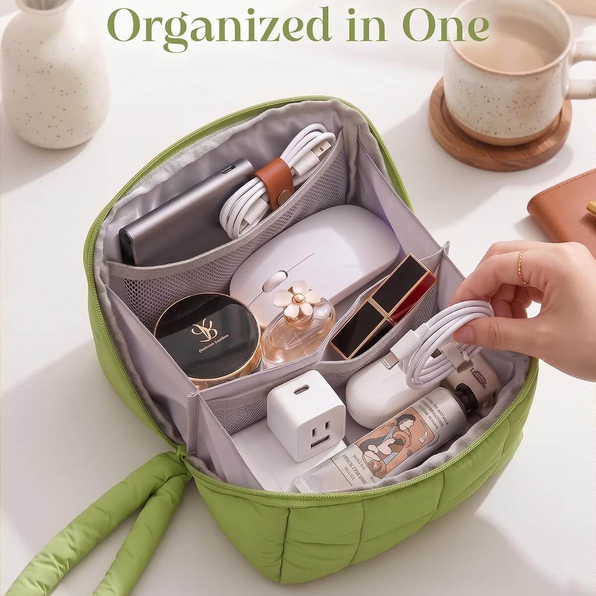 A green puffy tech pouch shown open on a white surface, filled with travel and beauty items. A hand is reaching toward the bag, and a coffee cup and vase are visible in the background.